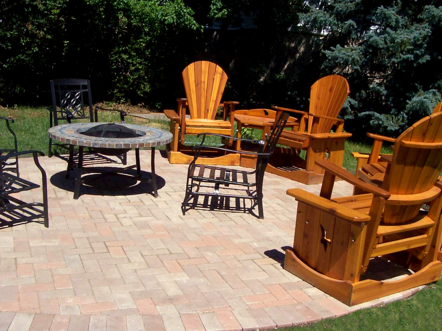 An outdoor patio area with wooden and metal chairs surrounding a round fire pit table on a brick paved surface