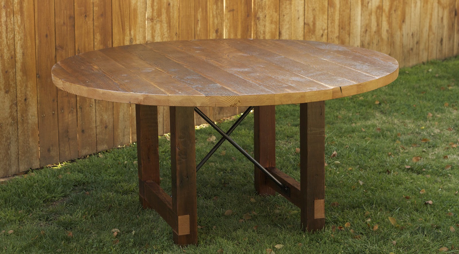 Hand-drawn ink illustration of a round rustic wooden table with a metal cross-brace base standing on grass in front of a wooden fence, warm brown and muted green tones.