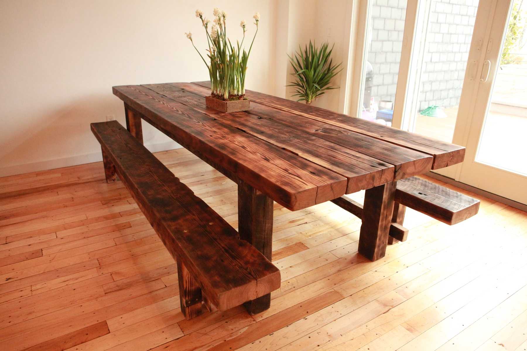 A long rustic wooden dining table with matching benches in a bright room with plants and glass doors, warm brown tones